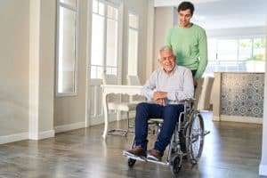 In this image, a man is pushing a man in a wheelchair down a hallway. The woman is smiling and appears to be happy. The man is wearing a green shirt and is pushing the wheelchair with both hands. The hallway is lined with hardwood floors and there are doors on either side. There is a window at the end of the hallway that lets in a lot of natural light. The overall atmosphere of the image is warm.