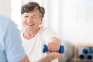 An older woman is shown standing next to a man who is holding a pair of dumbbells. The woman is smiling and appears to be in good spirits. The man is also smiling and appears to be helping the woman with her exercise routine.